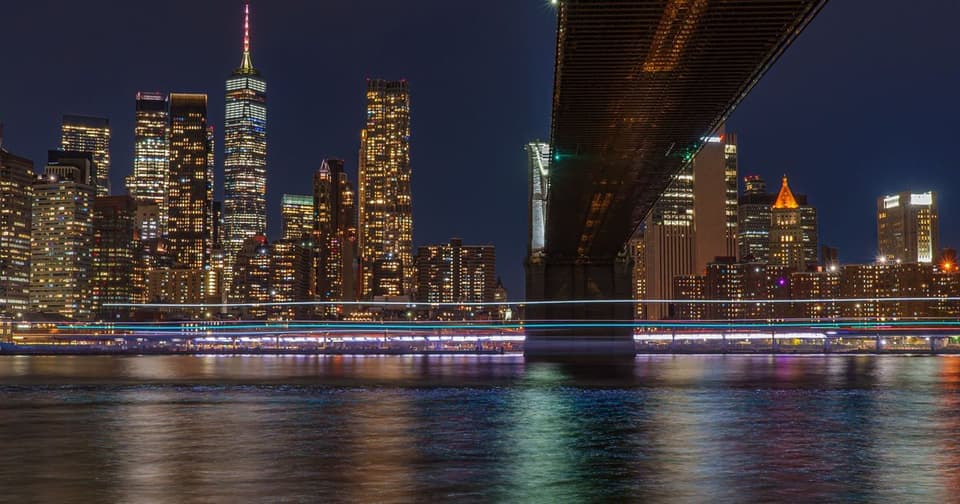Skyscrapers with night lights reflected in the water and the dark underside of a bridge in the foreground