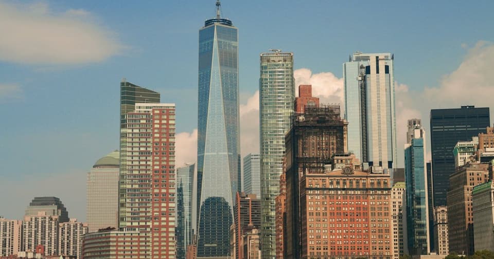 A glass skyscraper with a pointed top rising against a blue sky and white clouds, and its surroundings