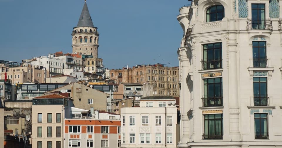 Süslü beyaz cepheli tarihi bir binanın yanında eski yapıların ardında yükselen Galata Kulesi