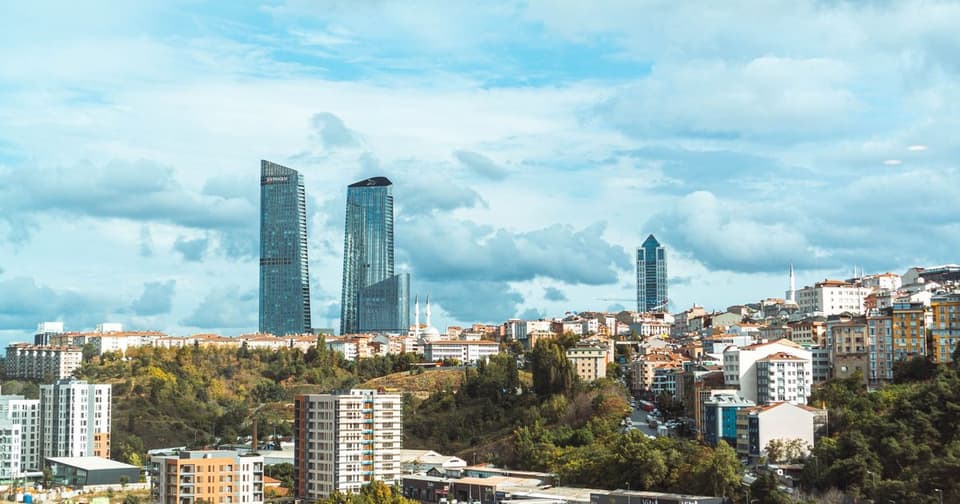 2 glass skyscrapers rising behind a neighborhood on a green hillside under a cloudy sky